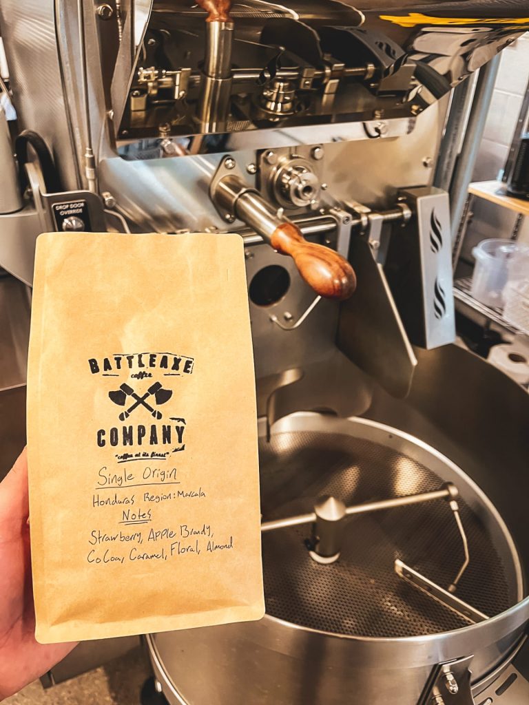 A hand holds a bag of BattleAxe coffee with a coffee roasting machine in the background. 